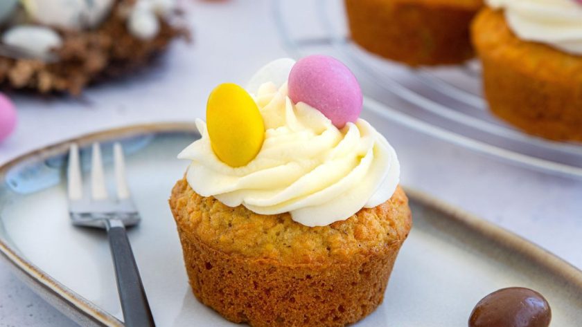 Babeczka z kremem waniliowym udekorowana żółtym i różowym jajeczkiem cukrowym; w tle widoczne inne słodycze na święta oraz gniazdko z czekoladowymi jajkami
