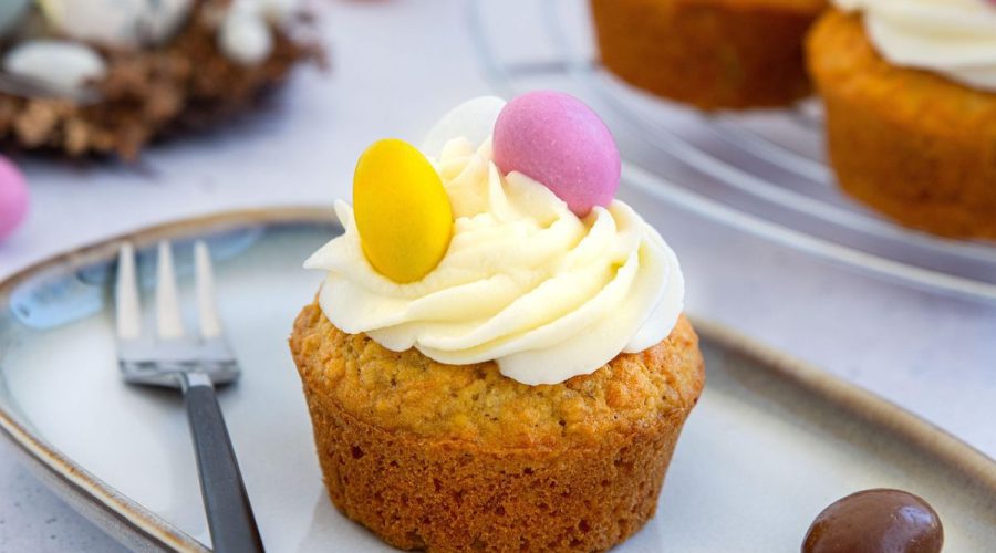 Babeczka z kremem waniliowym udekorowana żółtym i różowym jajeczkiem cukrowym; w tle widoczne inne słodycze na święta oraz gniazdko z czekoladowymi jajkami