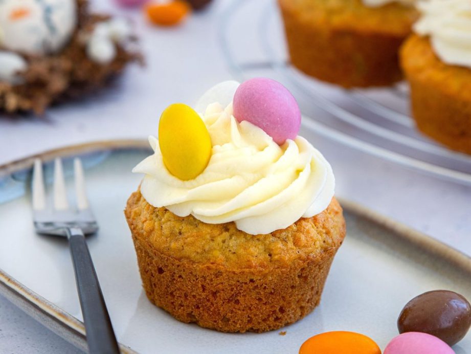 Babeczka z kremem waniliowym udekorowana żółtym i różowym jajeczkiem cukrowym; w tle widoczne inne słodycze na święta oraz gniazdko z czekoladowymi jajkami